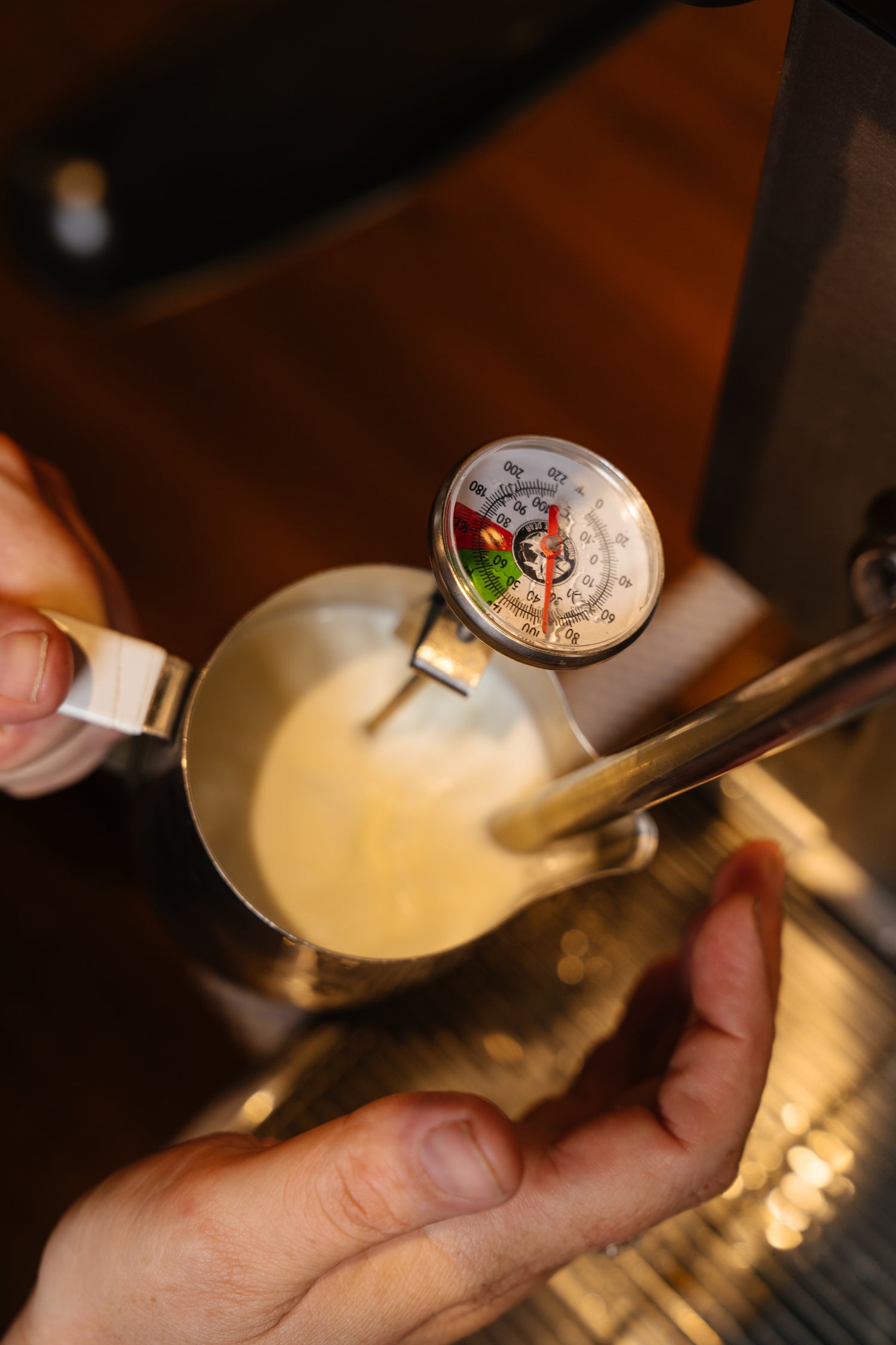 Hands holding a metal pitcher with a milk thermometer while steaming milk