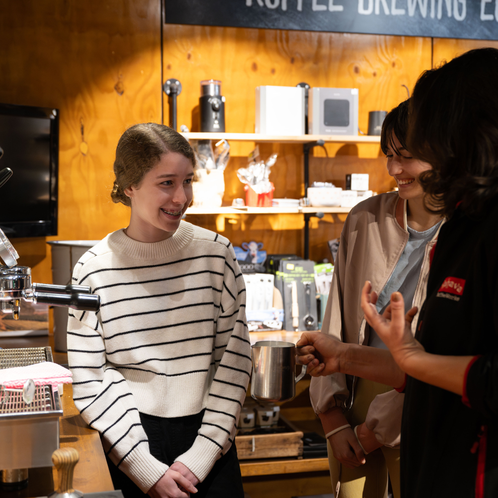 A woman in a cream sweater with thin black horizontal stripes stands smiling in a coffee shop setting, facing another person holding a metal pitcher. Shelves with coffee equipment and a wood-paneled wall are visible in the background.