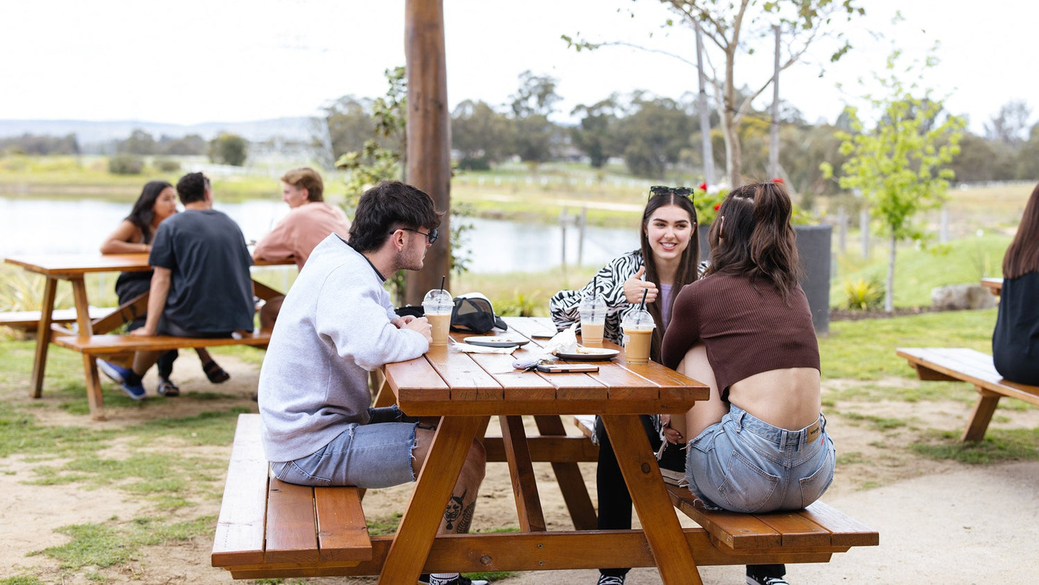 Group of young adults sitting at outdoor wooden picnic tables with iced coffees