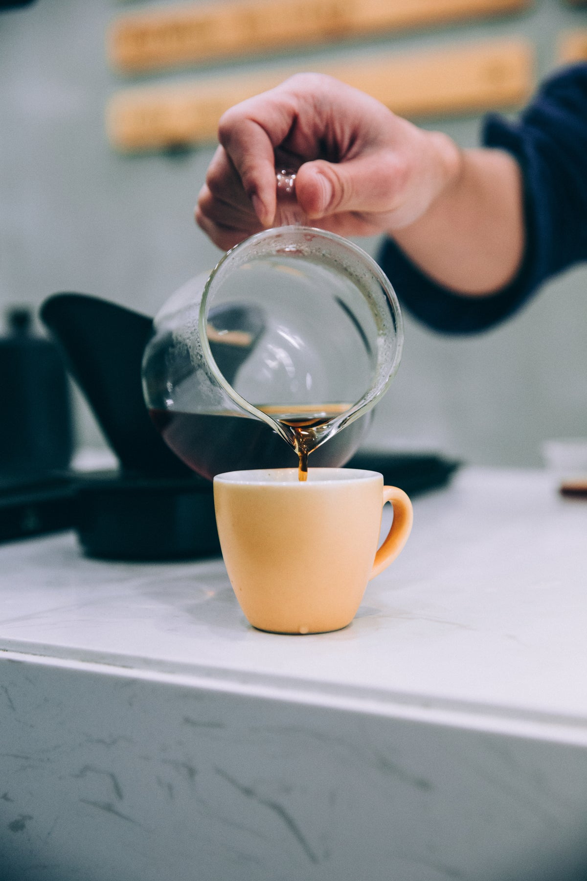 Hand pouring brewed coffee from a glass carafe into a small yellow ceramic cup